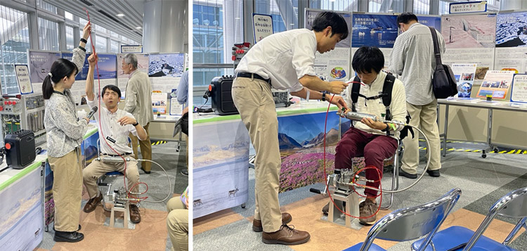 手動大気採取装置(MSE)実演中の藤田さん(左)と体験中の来場者。手動ポンプのハンドルをひたすら回して、加圧した金属容器に大気を採取する。藤田さんはシカゴ-羽田便13時間半のフライトで12本のフラスコに大気を充填し続けた(国立極地研究所提供)